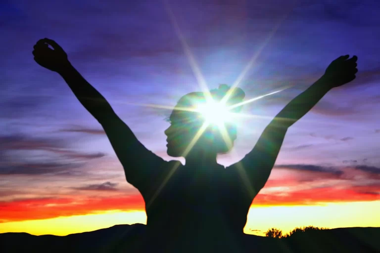 A silhouette of a woman raising her hands and light reflects her head